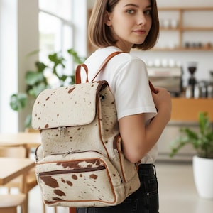 Puede incluir: Mochila con estampado de piel de vaca en crema y marrón. La bolsa tiene un cierre de solapa, un bolsillo delantero y correas de cuero marrón. La mochila se está usando.