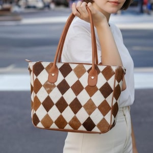 Puede incluir: Un bolso tote con un patrón de diamantes en tonos marrón, blanco y beige. El bolso tiene asas y ribetes de cuero marrón. El bolso está siendo sujetado.
