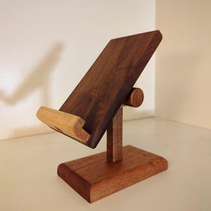 May include: A wooden tablet stand with a dark brown angled back and a lighter brown cradle. The stand is supported by a vertical post and a rectangular base, all made of wood. The design is simple and functional.