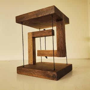 Può includere: Una scultura in legno dal design geometrico. La scultura presenta una base e una parte superiore marrone scuro, con elementi verticali e orizzontali marrone chiaro sospesi da corde nere. Il design crea un'illusione ottica.