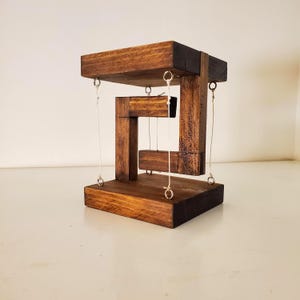 May include: A wooden sculpture with a dark brown finish. The sculpture has a rectangular base and top, connected by vertical supports. A central, floating wooden element is suspended by thin silver-colored wires, creating an optical illusion. The overall design is geometric and modern.