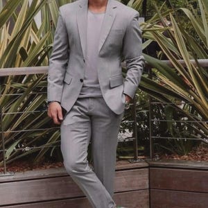 Può includere: Un completo grigio chiaro, composto da giacca e pantaloni, abbinato a una t-shirt grigio chiaro e scarpe da ginnastica bianche. L'outfit è completato da occhiali da sole e un orologio. Lo sfondo presenta piante verdi e una staccionata di legno.