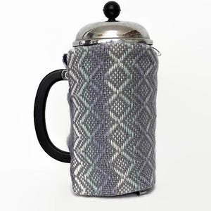 May include: A French press coffee maker with a black handle and a silver lid. The glass carafe is wrapped in a woven fabric cosy with a diamond pattern in shades of grey, blue, and white. The cosy is designed to keep the coffee warm.