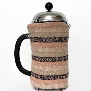 Puede incluir: Una cafetera de émbolo con una funda de tela tejida. La funda presenta rayas horizontales en tonos rosa, beige y negro. La cafetera tiene una tapa de acero inoxidable y un asa negra.