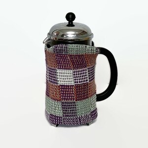Pode incluir: Uma cafeteira de prensa francesa de aço inoxidável com uma capa de tecido xadrez em tons de roxo, laranja e branco.