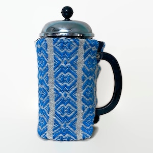Puede incluir: Una cafetera de prensa francesa plateada con una cubierta de tela azul y blanca con estampado.