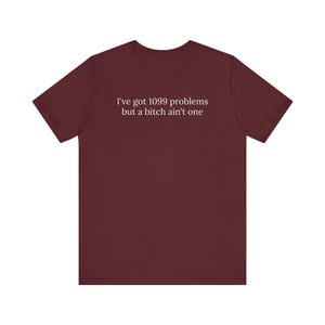 May include: A maroon t-shirt with the white text "I've got 1099 problems but a bitch ain't one". The top has short sleeves and a round neck.