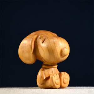 Pode incluir: Uma estatueta de madeira de um c&atilde;o de desenho animado, esculpida em madeira de cor clara. O c&atilde;o est&aacute; sentado com as patas juntas e os olhos fechados. A estatueta tem orelhas grandes e uma cabe&ccedil;a redonda, com gr&atilde;o de madeira vis&iacute;vel.