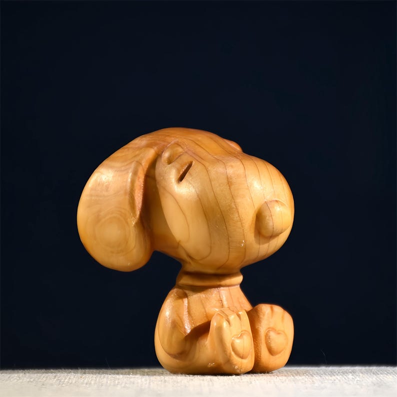 Pode incluir: Uma estatueta de madeira de um c&atilde;o de desenho animado, esculpida em madeira de cor clara. O c&atilde;o est&aacute; sentado com as patas juntas e os olhos fechados. A estatueta tem orelhas grandes e uma cabe&ccedil;a redonda, com gr&atilde;o de madeira vis&iacute;vel.