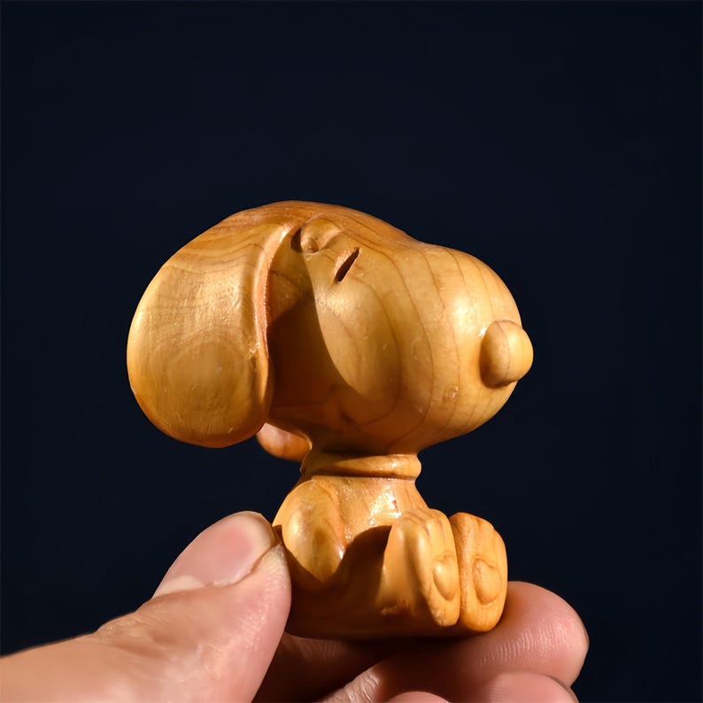 Pode incluir: Estatueta de madeira de um c&atilde;o de desenho animado, esculpida em madeira de cor clara. O c&atilde;o est&aacute; sentado com a cabe&ccedil;a ligeiramente inclinada. A estatueta tem um acabamento liso e &eacute; segurada em uma m&atilde;o sobre um fundo escuro.