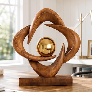 Puede incluir: Una escultura decorativa con una esfera dorada pulida suspendida dentro de un marco curvo con vetas de madera marrón. La estructura descansa sobre una base rectangular de madera, mostrando líneas fluidas que crean una forma geométrica abierta.