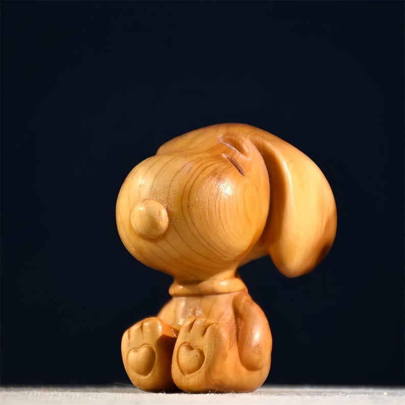 Pode incluir: Estatueta de madeira castanho-claro de um c&atilde;o de desenho animado. O c&atilde;o est&aacute; sentado com a cabe&ccedil;a apoiada nas patas. A estatueta tem detalhes em forma de cora&ccedil;&atilde;o nas patas, sobre um fundo escuro.