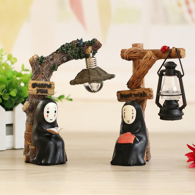 May include: Two decorative figurines featuring characters from a popular animated film. One figure is reading under a lamp, the other is holding a fan under a lantern. The figurines are black with white masks and are set on a wooden base.