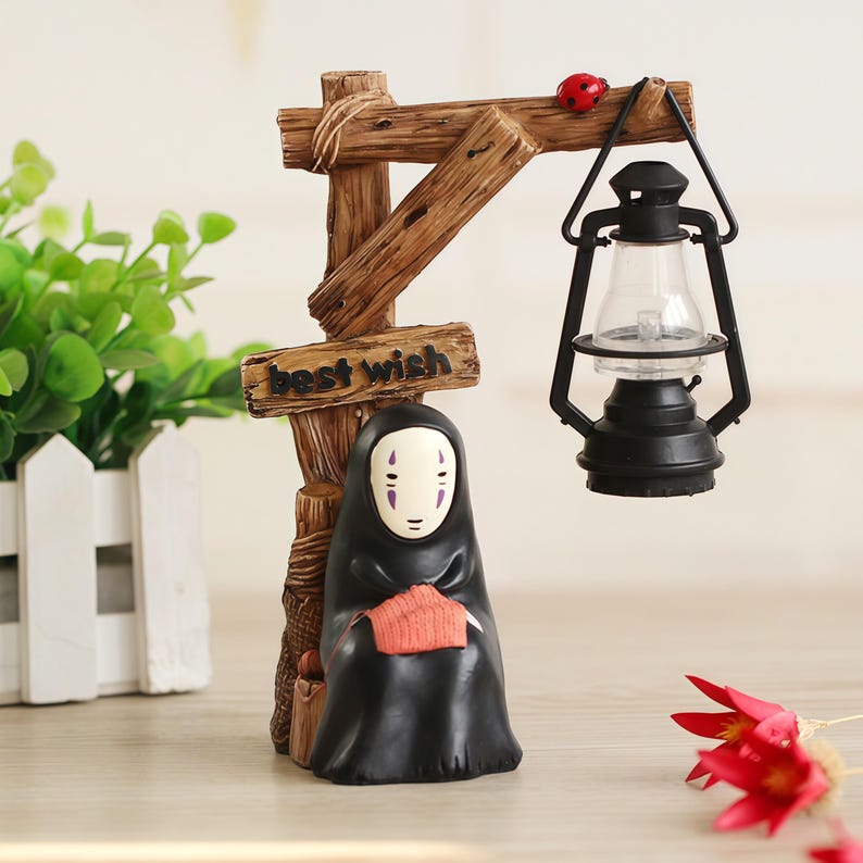 May include: A decorative figurine featuring a character from Spirited Away, with a black cloak and a white mask. The figurine is holding a small orange item. A wooden sign with the words "best wish" is attached to a wooden post, and a black lantern hangs from the post.