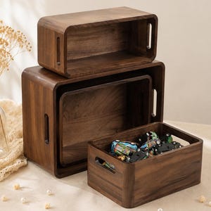 Puede incluir: Un conjunto de tres cajas de almacenamiento rectangulares de madera marrón oscuro con esquinas redondeadas y asas laterales integradas. Las cajas están apiladas, con la caja más pequeña al frente conteniendo varios coches de juguete coloridos.
