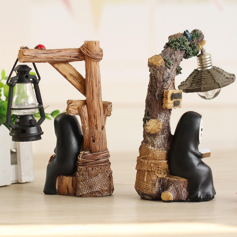 May include: Two decorative bookends. One features a black figure seated beneath a wooden structure with a lantern. The other bookend has a black figure seated beneath a tree with a lamp. Both figures have a white mask.