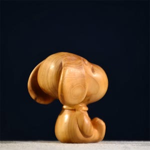 Pode incluir: Uma estatueta de madeira de um c&atilde;o, esculpida em madeira de cor clara. O c&atilde;o est&aacute; sentado com a cabe&ccedil;a virada para o lado, mostrando orelhas grandes e ca&iacute;das. A estatueta tem um acabamento liso e polido e est&aacute; sobre um fundo escuro.