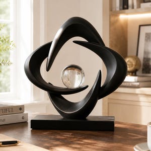 Puede incluir: Una escultura abstracta de color negro mate con formas curvas entrelazadas que crean una abertura central. Una esfera de cristal transparente está situada en el centro de la estructura negra, que descansa sobre una base rectangular negra.