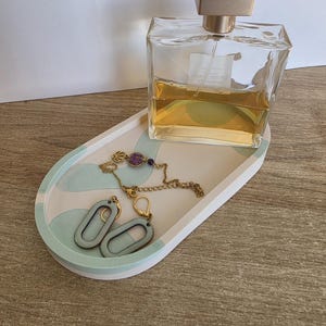 Peut inclure: Un plateau décoratif ovale avec une bordure blanche et des accents bleu clair et pêche. Le plateau contient un flacon de parfum, un collier chaîne doré avec une pierre violette et une paire de boucles d'oreilles ovales bleu clair.