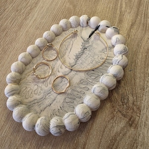 Peut inclure: Un plateau à bijoux ovale avec un motif marbré et une bordure perlée. Le plateau contient plusieurs bagues dorées et une boucle d'oreille créole dorée. Les perles sont blanches avec des marbrures grises. Le plateau est posé sur une surface en bois.