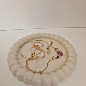 Peut inclure: Un plateau à bijoux blanc effet marbre avec un bord festonné, contenant des bagues, des bracelets et un collier dorés avec un charm violet. Le plateau est rond et posé sur une surface blanche.