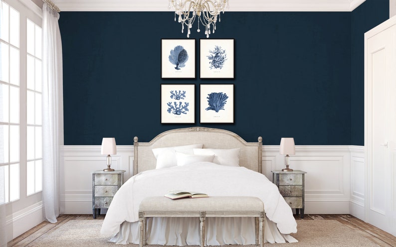 Op de afbeelding: Een wit bed met een wit hoofdeinde en een wit dekbed. Het bed staat in een kamer met donkerblauwe muren en witte lijstwerk. Er hangen vier ingelijste prints aan de muur boven het bed, elk met een blauwe en witte illustratie van een koraal. De prints zijn in een vierkant gerangschikt.