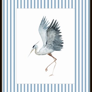 May include: A watercolor painting of a gray heron with its wings spread, standing on one leg, against a white background. The heron is depicted in a light blue frame with white stripes.