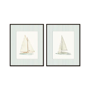 May include: Two framed watercolor prints of sailboats. Each print features a white sailboat with a beige sail on a light blue and white striped background. The frames are black.