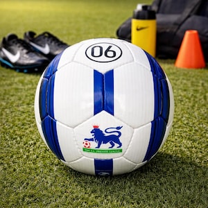 May include: A white and blue football with the Premier League logo, number 06, and blue stripes. Black football boots and a yellow water bottle are in the background on a green pitch.