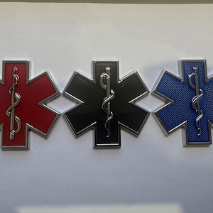 Puede incluir: Tres emblemas metálicos de la Estrella de la Vida en rojo, negro y azul. Cada emblema tiene un borde plateado y un bastón de Asclepio plateado en el centro. Los emblemas rojo y azul tienen una superficie texturizada.