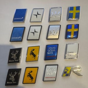 Peut inclure: Une collection d'emblèmes de voiture rectangulaires avec divers motifs et textes. Les emblèmes incluent le logo Polestar, une couronne, une croix, un élan et le texte "VOLVO OCEAN RACE". Les couleurs incluent le bleu, le blanc, le jaune et l'argent.
