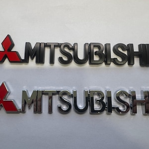 Puede incluir: Dos emblemas de coche Mitsubishi. Uno es negro con un logotipo rojo, el otro es cromado con un logotipo rojo. La palabra "MITSUBISHI" está escrita en mayúsculas en ambos emblemas. El logotipo de Mitsubishi es una forma de diamante rojo.