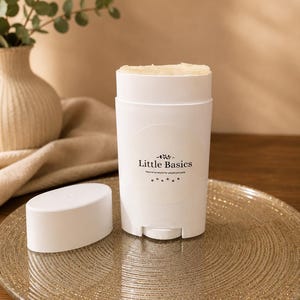 May include: A white deodorant stick with the text "Little Basics" and a matching cap sits on a gold-colored plate. The product is labeled "Natural products for people and pets". A vase with greenery is in the background.