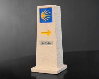 May include: A beige, textured pillar with a blue square and yellow shell design at the top. A yellow arrow points right below the blue square. The text "Km 772,000" is displayed on a small white rectangle.