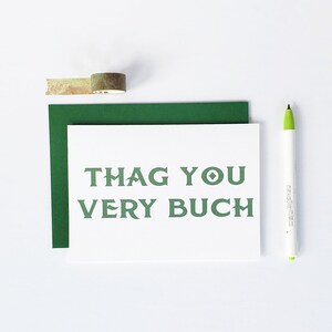 May include: A white card with green text that reads "THAG YOU VERY BUCH". The card is on top of a green envelope. There is a roll of tape and a green marker on a white surface.