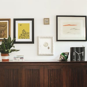 May include: A dark wood cabinet with two doors and a white wall with framed artwork, including a black and white print of birds flying over a yellow background, a black and white print of a landscape, and a framed print of a seascape. There is a green glass decorative object and a clock made of three books on top of the cabinet.