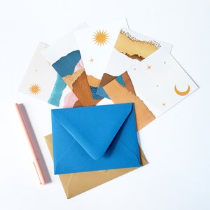 May include: A set of six blank cards with a gold and brown abstract design. The cards are stacked on top of a blue envelope and a gold envelope. A pink pen is lying on the white surface.