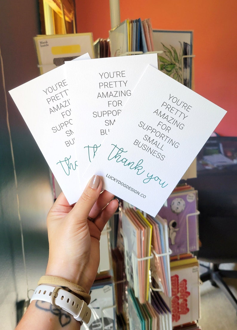 May include: Three white cards with green text that reads "You're pretty amazing for supporting small business. Thank you." The cards are stacked on top of each other and held in a hand. The text on the bottom card reads "LuckyDogDesign.co".