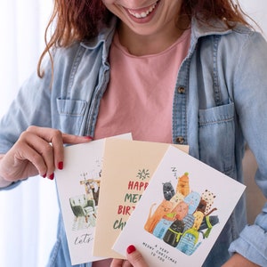 May include: A woman holds three greeting cards. One card features a group of cats with the text "A Very Meowy Christmas to You". Another card has the text "Happy Meow Birthday to You". The third card has a cat sitting in a chair with the text "Let's Get Cozy".