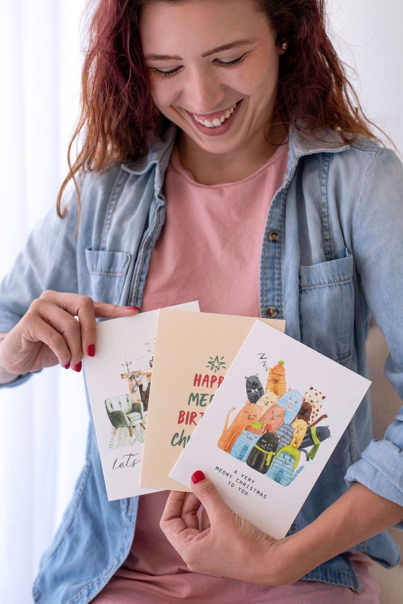 May include: A woman holds three greeting cards. One card features a group of cats with the text "A Very Meowy Christmas to You". Another card has the text "Happy Meow Birthday to You". The third card has a cat sitting in a chair with the text "Let's Get Cozy".