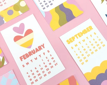 May include: A collection of colorful monthly calendars with unique designs. Each calendar features a different month, such as February, April, and August, with a playful font and vibrant color schemes. The calendars are arranged on a pink surface.