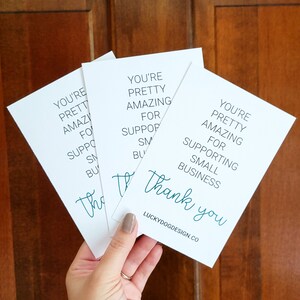 May include: Three white cards with teal text that reads "You're pretty amazing for supporting small business. Thank you." The cards are stacked on top of each other and held in a hand. The text "LUCKYDOGDESIGN.CO" is printed on the bottom of the card.