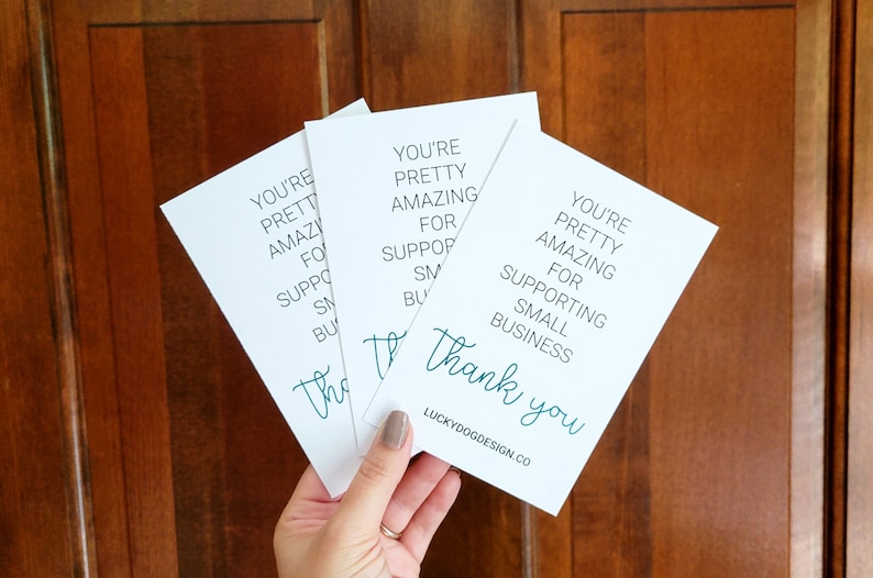 May include: Three white cards with teal text that reads "You're pretty amazing for supporting small business. Thank you." The cards are stacked on top of each other and held in a hand. The text "LUCKYDOGDESIGN.CO" is printed on the bottom of the card.