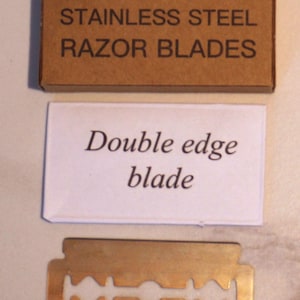 May include: A brown cardboard box labeled "x5 PREMIUM STAINLESS STEEL RAZOR BLADES" sits above a white card that reads "Double edge blade." A gold-colored double edge razor blade is at the bottom.