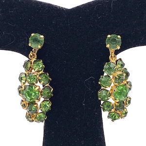 May include: A pair of vintage gold-toned earrings with clusters of green rhinestones. Each earring features a large, round green rhinestone at the top, with a cascade of smaller green rhinestones below. The earrings are displayed on a black surface.