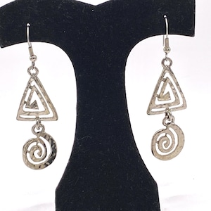 May include: Silver-toned earrings with a geometric design. Each earring features a triangle shape with an open center, connected to a spiral shape below. The earrings are displayed on a black stand against a white background.