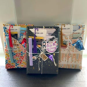 May include: Three rectangular gift packages wrapped in patterned paper, each containing a reading list card, a bookmark with a tassel, a highlighter, and small chocolate squares. The center package is black with gold stars and a sticker reading READ SOMETHING TODAY.
