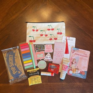 May include: A white canvas zippered pouch with a red cherry pattern, accompanied by a Pop-Secret popcorn bag, colorful sticky tabs, two pens, various stickers, a reading list bookmark, a tea bag, lip balm, chocolate, hand cream, and a pomegranate face mask.