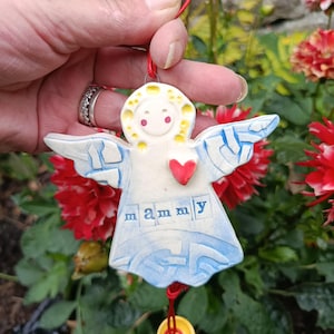 Peut inclure: Un ornement d'ange en céramique avec un corps blanc aux accents bleus, un cœur rouge et le texte "mammy" imprimé sur le devant. L'ornement est suspendu par un cordon rouge avec un petit bouton jaune attaché au bas de la figurine.