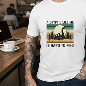 Pode incluir: Camiseta branca com um design retrô. O texto diz "A Cryptid Like Me Is Hard To Find". O gráfico mostra uma criatura críptida em um lago, com um sol e árvores. O design tem um estilo vintage, que lembra as aventuras ao ar livre.